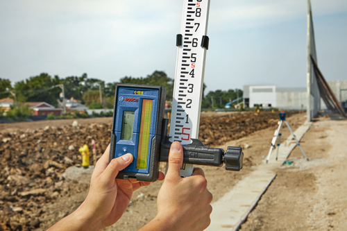 Bosch leveling device and measuring stick held by hands, outdoors at a construction site.