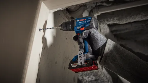 Bosch GSB18V-490 drill is used to drill a hole in a white wooden surface. The drill is held by a gloved hand.