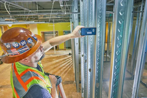 Man holding a leveling tool, wearing a hard hat with "www" and American flag stickers, and an orange and neon green safety vest.