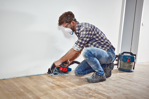 A person wearing safety glasses and a mask cuts a wooden floor with a Bosch circular saw. Bosch Professional vacuum cleaner nearby.