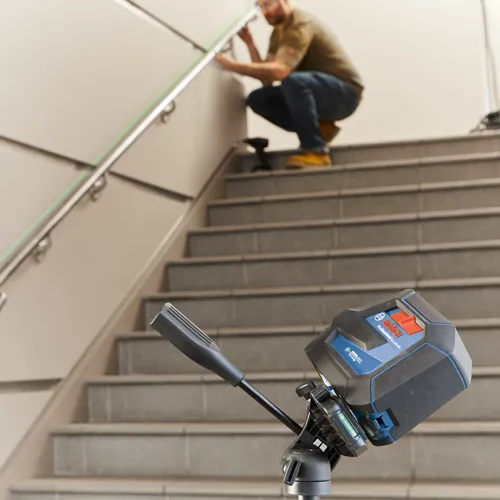 Bosch Professional GLL 100-40G laser level on tripod, with a person squatting on the stairs adjusting the handrail.