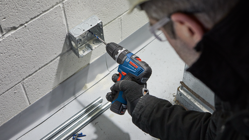 Bosch GSB12V-300 HD cordless drill being used to drill into a metal electrical box mounted on a concrete wall. A person wearing safety glasses and a hard hat is using the drill. Blue and silver pipes are on a surface below.