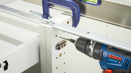 Bosch GSB12V-300 HD drill drilling into white cabinetry; metal clamps, level are also visible.