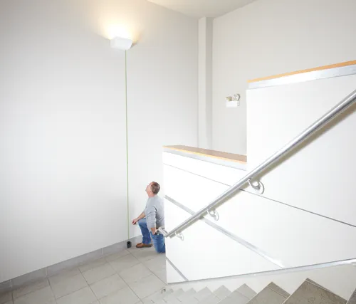 A person kneels near a wall, using a measuring device. The device is connected to a light fixture on the wall. A staircase and a handrail are also visible.
