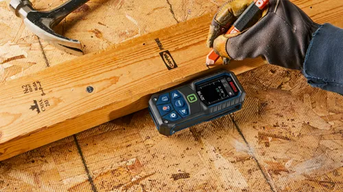 Bosch BLAZE laser measure, visible text reads "2x 2’08”", "5’05 5/16", and a gloved hand with a pencil is marking a wooden board with markings.