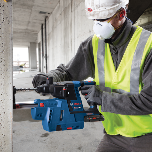 Person wearing a hard hat, face mask, and high-visibility vest using a Bosch GBH18V-26 BULLDOG rotary hammer drill to drill into a concrete pillar.