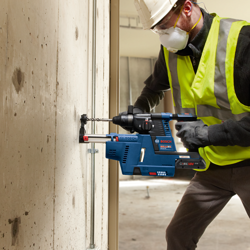 Worker using a Bosch GBH18V-26 Bulldog rotary hammer drill with a CORE18V battery, wearing a hard hat, safety glasses, ear plugs, and a respirator mask.