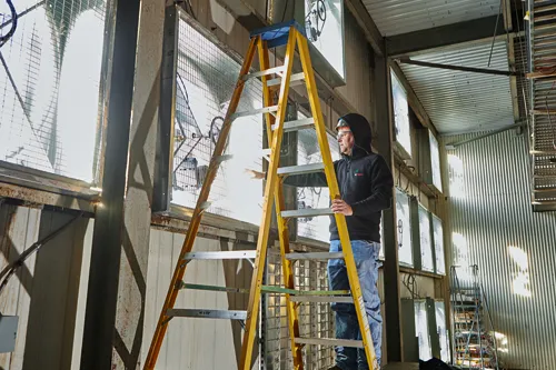 Person on a yellow ladder, wearing safety glasses and a black jacket with a green Bosch logo, in a building with large windows.
