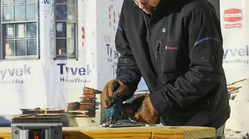 Person wearing a jacket with "BOSCH" logo using a jigsaw on lumber.