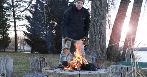 Person in winter clothes adds a log to a fire pit with burning flames; trees, stumps, and a lake are in the background.
