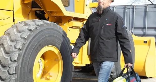 A person with a beard in a black jacket with a "Bosch" logo on it walks by a large yellow construction vehicle. The person is holding a bag with the "Bosch" logo.