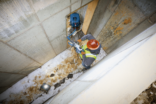 Person wearing a hard hat and safety vest uses a power tool on concrete wall; a vacuum cleaner is nearby.