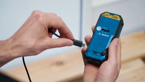 A person holding a blue and black Bosch device and a USB cable, with text "IEC 60825-1:2014 <1mW, 515 nm" and "LASER RADIATION DO NOT STARE INTO BEAM CLASS 2 LASER PRODUCT".