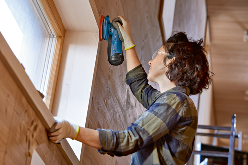 Person wearing safety glasses and gloves, working on a wooden window frame, using a Bosch GEX 12V-125 sander.