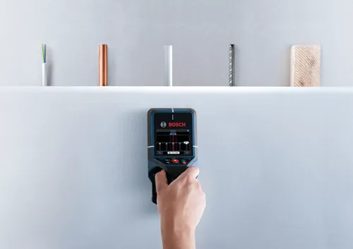 Hand holding a  Bosch wall scanner device; objects above include cable, copper pipe, plastic pipe, rebar, and wood.