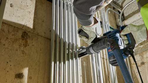 A person wearing gloves is using a Bosch RH328VC rotary hammer to install electrical conduit.