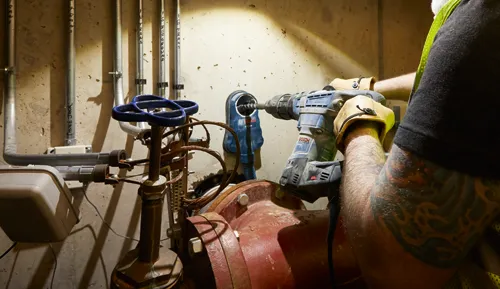 Bosch drill being used near pipes and valves, person wearing gloves, tattoos visible. Pipes labeled "TRANS", "PHONE JACK", "ISO GROUND".