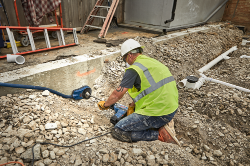 A person wearing a hard hat, safety glasses, a yellow reflective vest, and gloves kneels on rocky ground, using a Bosch tool to work on a construction project, other tools and materials visible.