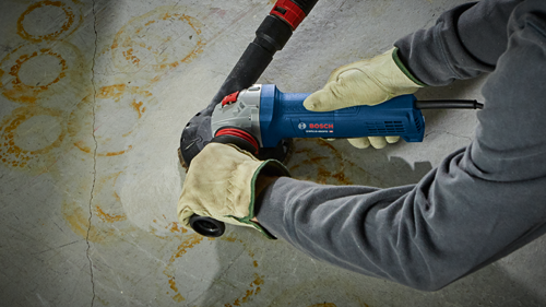 A person wearing gloves using a Bosch GWS10-45PD angle grinder, with a dust collection attachment against a concrete wall.