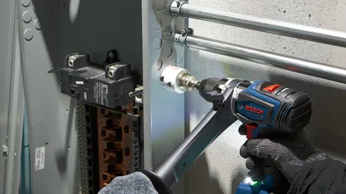 Bosch GSR18V-1330C drill with a hole saw drilling into a metal electrical box next to a 200A circuit breaker and conduit pipes, person wearing gloves.