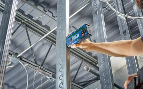 Bosch measuring tool, with digital display, being used against a metal stud. A person holds the tool.