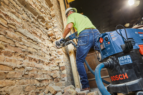 Person wearing safety gear operates a power tool on a brick wall, a HEPA Ready Bosch vacuum cleaner nearby.