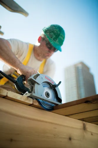 Man wearing safety glasses and a green hard hat using a Bosch CS 10 circular saw to cut wood.