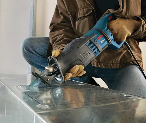 A person wearing work gloves, blue jeans, and a brown jacket uses a Bosch RS428 reciprocating saw to cut metal.