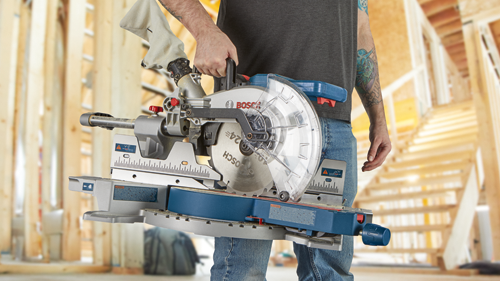 Bosch circular saw, held by a person with visible tattoos, set against a wooden structure backdrop.