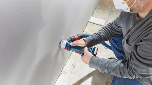 Man wearing mask using Bosch drywall cutout tool, model GLM18V-40, with dust collection hose attached.