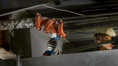 Bosch cordless impact wrench being used on pipes connected with Rigidlite FireLock fittings, a worker wearing safety glasses and a hard hat.
