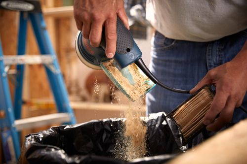 A person emptying a Bosch sander into a bag, dust is visible in the air. The sander is blue and black.