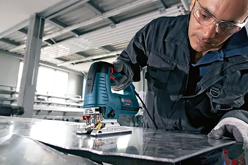 Person wearing safety glasses and gloves uses a Bosch jigsaw to cut metal.