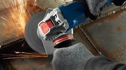 Angle grinder with sparks flying, being used by gloved hands. "X-Lock" is visible on the grinder.
