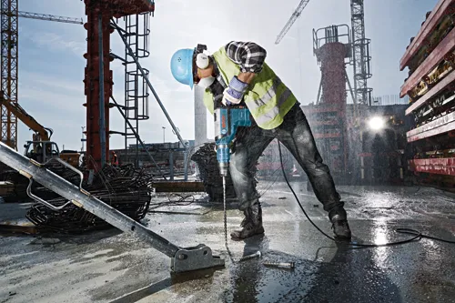 Person wearing a blue hard hat, ear protection, safety vest, and work gloves using a Bosch RH745 rotary hammer drill on a construction site.