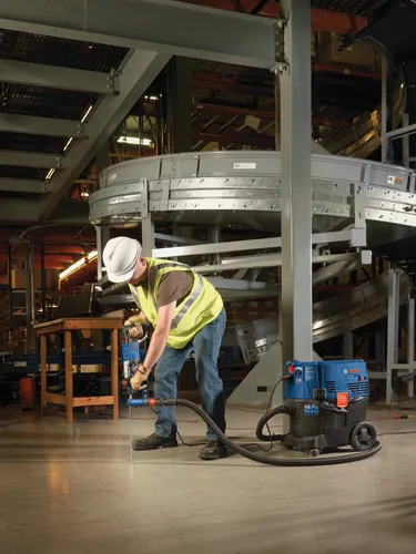Person wearing a hard hat, safety vest drilling into floor with a blue and black drill, next to a HEPA Ready vacuum.