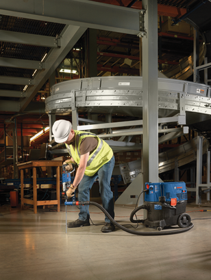 Person wearing a hard hat, safety vest drilling into floor with a blue and black drill, next to a HEPA Ready vacuum.
