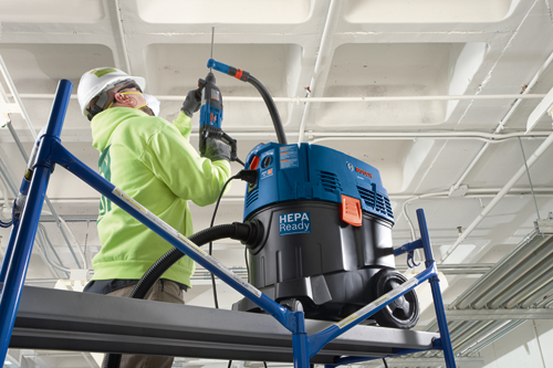 A person wearing safety gear, working on a ceiling, operating a Bosch drill and a Bosch VAC000A HEPA Ready vacuum cleaner.
