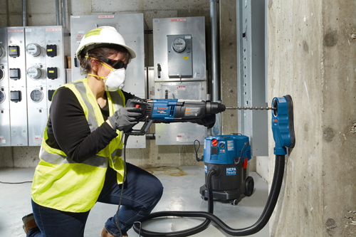 A person wearing safety glasses, a respirator, a white hard hat and a yellow reflective vest uses a Bosch BULLDOG Xtreme MAX rotary hammer drill to drill into a concrete wall. A Bosch HEPA Ready vacuum cleaner is on the floor with a hose connected to a dust collection shroud attached to the wall.
