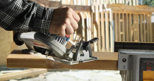 A person wearing a plaid shirt using a Bosch circular saw to cut a piece of wood.