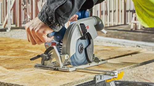 Bosch circular saw cutting plywood. The saw features the "BOSCH" brand name and the "GKS18V-22L" model code. A person wearing a black jacket is operating the saw.