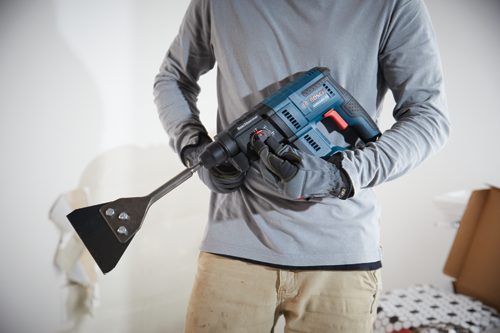 Person holding Bosch GBH18V-21 rotary hammer with a scraper attachment. The person wears gloves and a gray long-sleeved shirt.