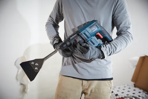 Person holding Bosch GBH18V-21 rotary hammer with a scraper attachment. The person wears gloves and a gray long-sleeved shirt.