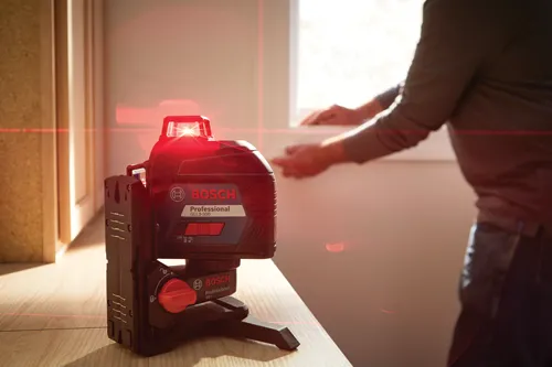 Bosch Professional GLL3-300 laser level, red laser lines projected onto wall, and person with arms visible by window.