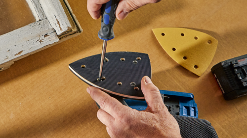 Person using a screwdriver to attach a sanding pad to a sander. The sander base and a spare pad are on a brown surface. A small piece of a white-painted wooden frame is in the background.