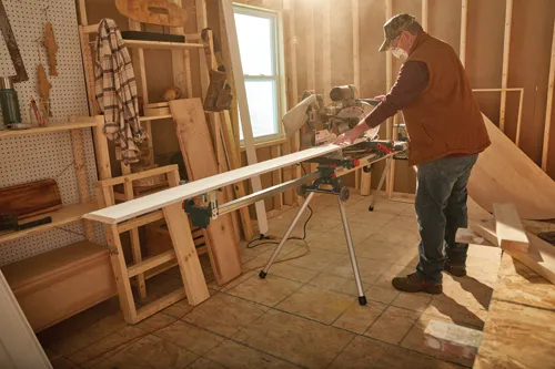 Man wearing mask and cap using a Bosch miter saw to cut a long, white board in a workshop setting.
