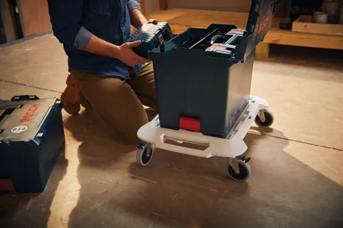 Man kneeling, holding a Bosch toolbox, with other Bosch toolboxes on a wheeled platform.