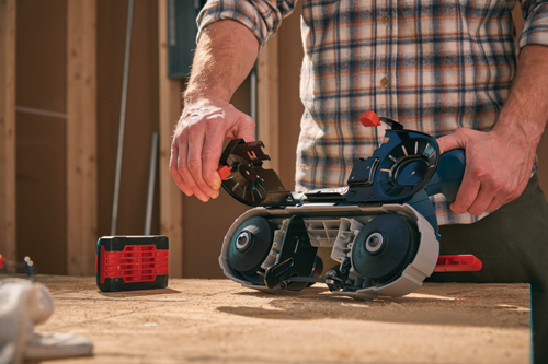 Person holding a band saw with black and red accents; a red battery pack lies on the wood surface.