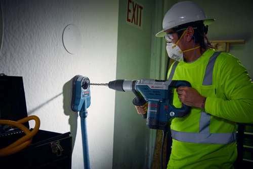 Person in safety attire using Boschhammer 11264EVS hammer drill. An additional Bosch HGC200 dust collection attachment is also mounted on the wall.