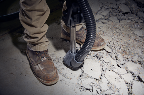 Person wearing brown boots and work pants, using a tool to break up concrete floor.